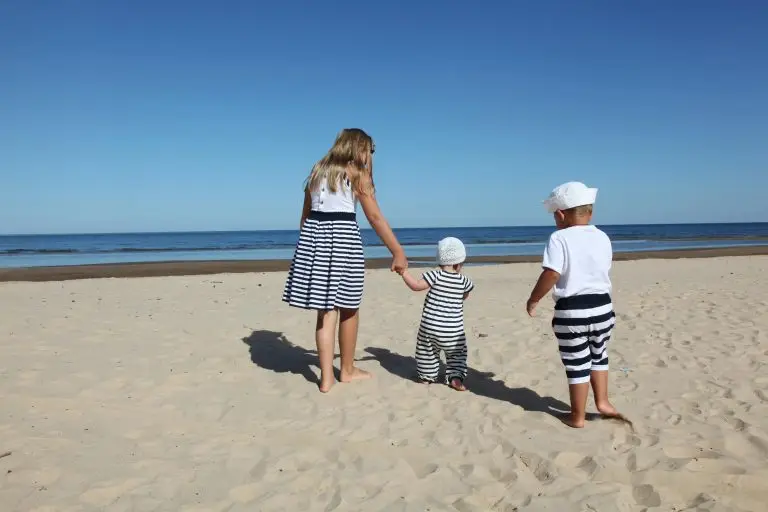 Three children in striped clothing walking on a sandy beach under a clear blue sky.